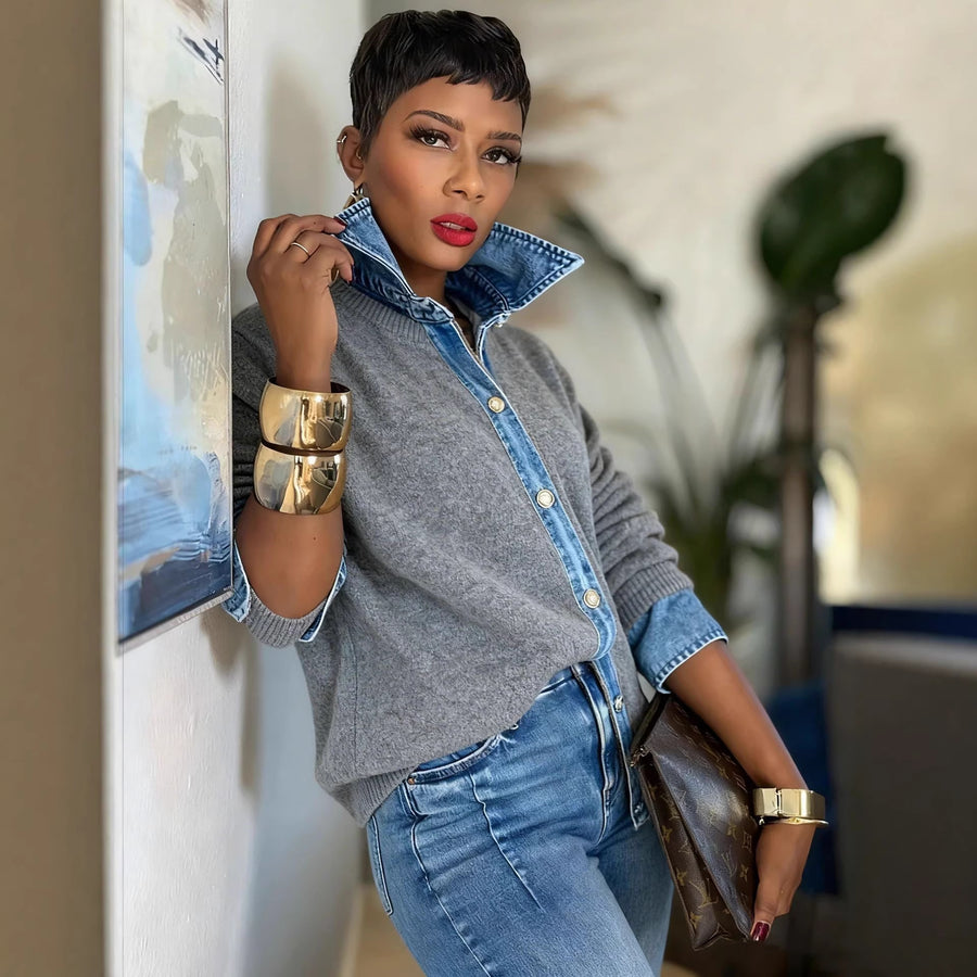 A woman wearing a grey knitted jumper finished with a denim shirt collar, a denim button placket and denim cuffs, giving the look of a shirt layered under a sweater, standing beside a framed picture.