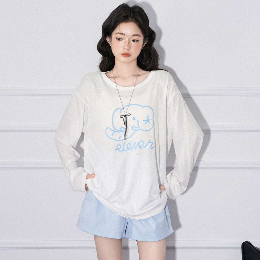A woman wearing a loose cream long sleeved top printed at the chest with a pale blue line-drawn elephant above the hand-lettered word eleven, with pale blue shorts, both hands pushed into pockets.
