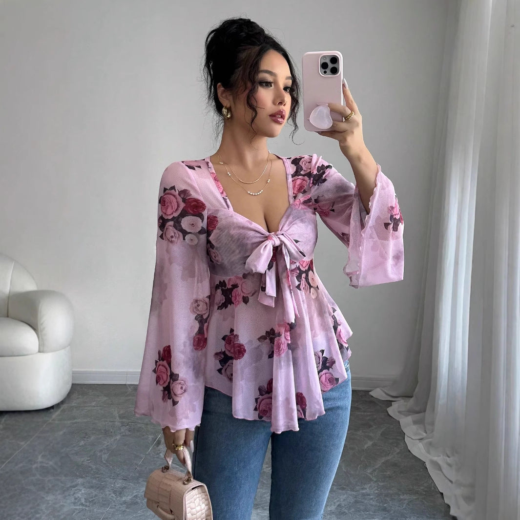 A woman wearing a lilac sheer chiffon top printed with pink and burgundy roses, tied in a bow at the bust above a soft peplum, with long wide flared sleeves, photographed in a mirror in a bare room.