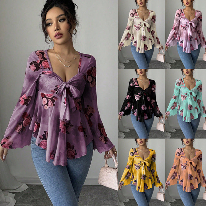 A woman wearing a lilac sheer chiffon top printed with pink and burgundy roses, tied in a bow at the bust above a soft peplum, with long wide flared sleeves, photographed in a mirror in a bare room.