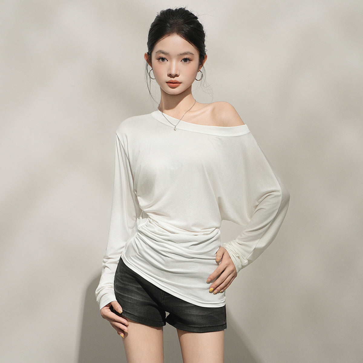 A woman wearing a cream long sleeved top with a wide boat neckline slipping off one shoulder, the soft fabric gathering at the waist and the sleeves cut full to the cuff.