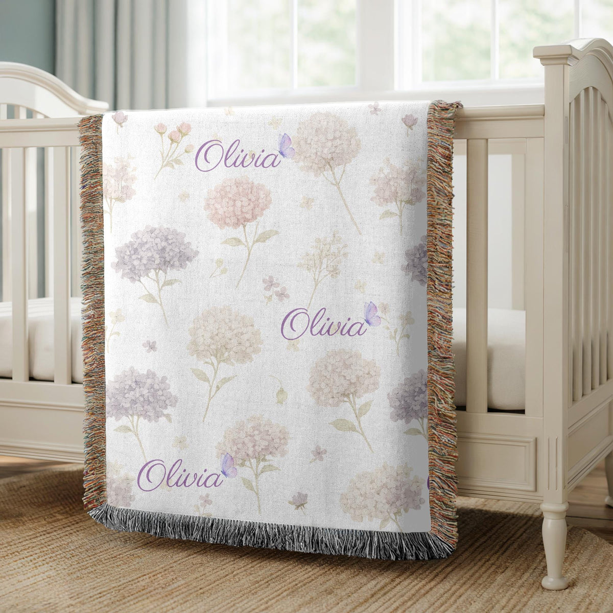 White fringed woven blanket draped over a cream crib, printed with soft blush and lilac hydrangeas and small purple butterflies, with the name Olivia repeated in purple script.