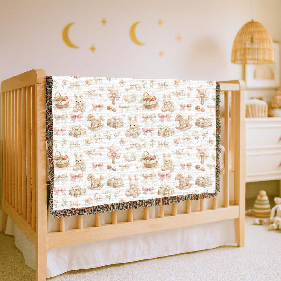White fringed woven blanket draped over a wooden crib, printed with watercolour bunnies, rocking horses, pink and cream bows, rattles, daisies and baskets of strawberries.