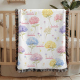 White fringed woven blanket over a wooden crib, scattered with pink, blue, yellow and purple watercolour hydrangeas, with the name Isabella repeated across it in pink script.