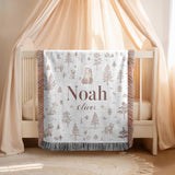 White fringed woven blanket over a cream crib, printed with line drawn pine trees, a bear and foxes in soft brown, with the name Noah in large serif and Oliver in script beneath.