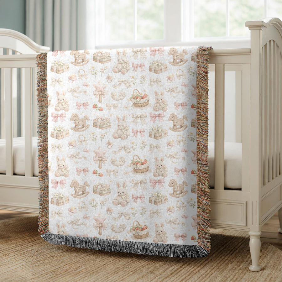 White fringed woven blanket draped over a wooden crib, printed with watercolour bunnies, rocking horses, pink and cream bows, rattles, daisies and baskets of strawberries.