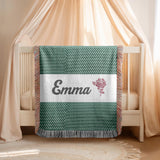 Green fringed woven blanket patterned with small cream dashes, crossed by a white band carrying the name Emma in dark script beside a single mauve rose and leaf drawn in outline.