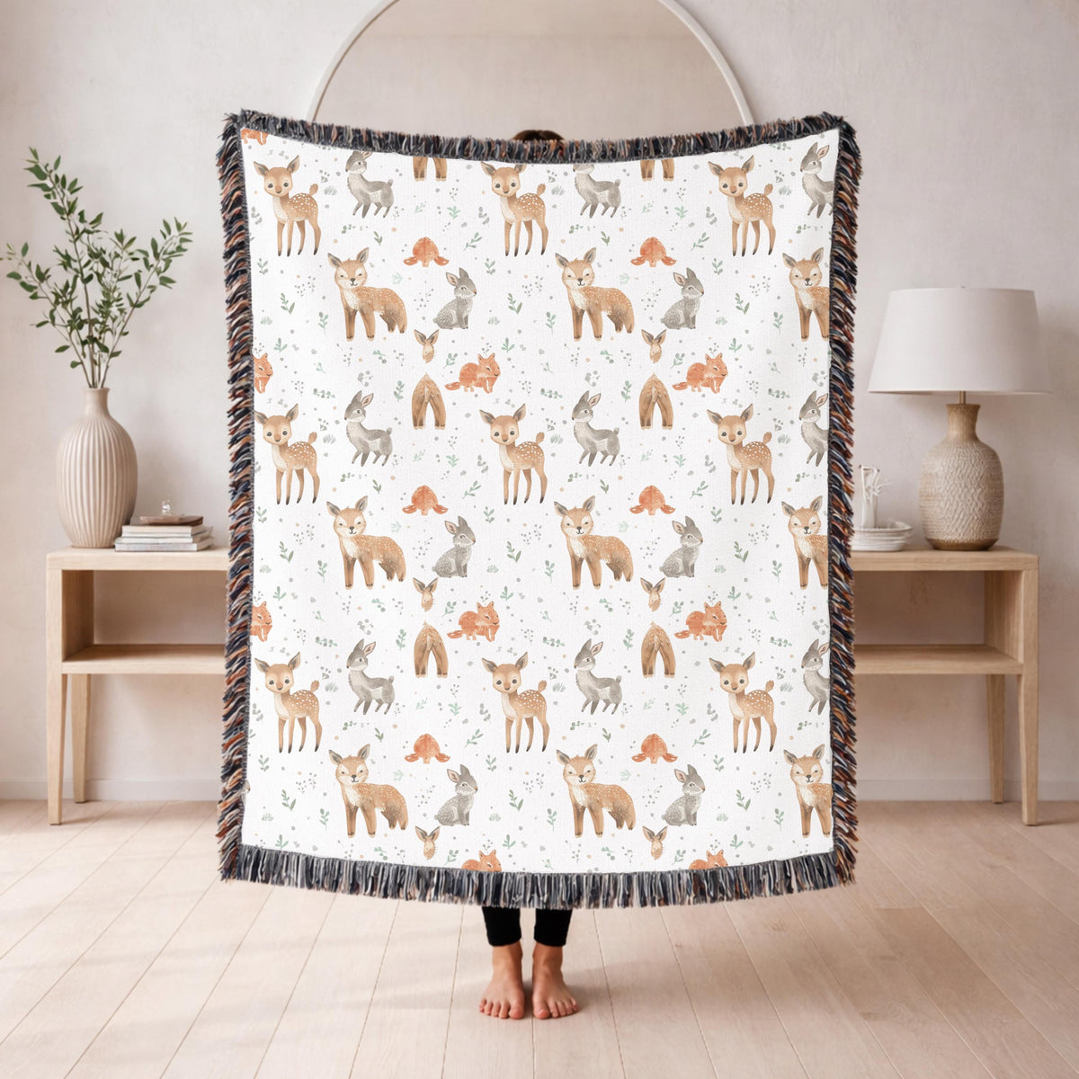 Fringed woven baby blanket held up in a living room, patterned all over with watercolour spotted fawns, foxes and grey rabbits among small orange mushrooms and pale green leaves.
