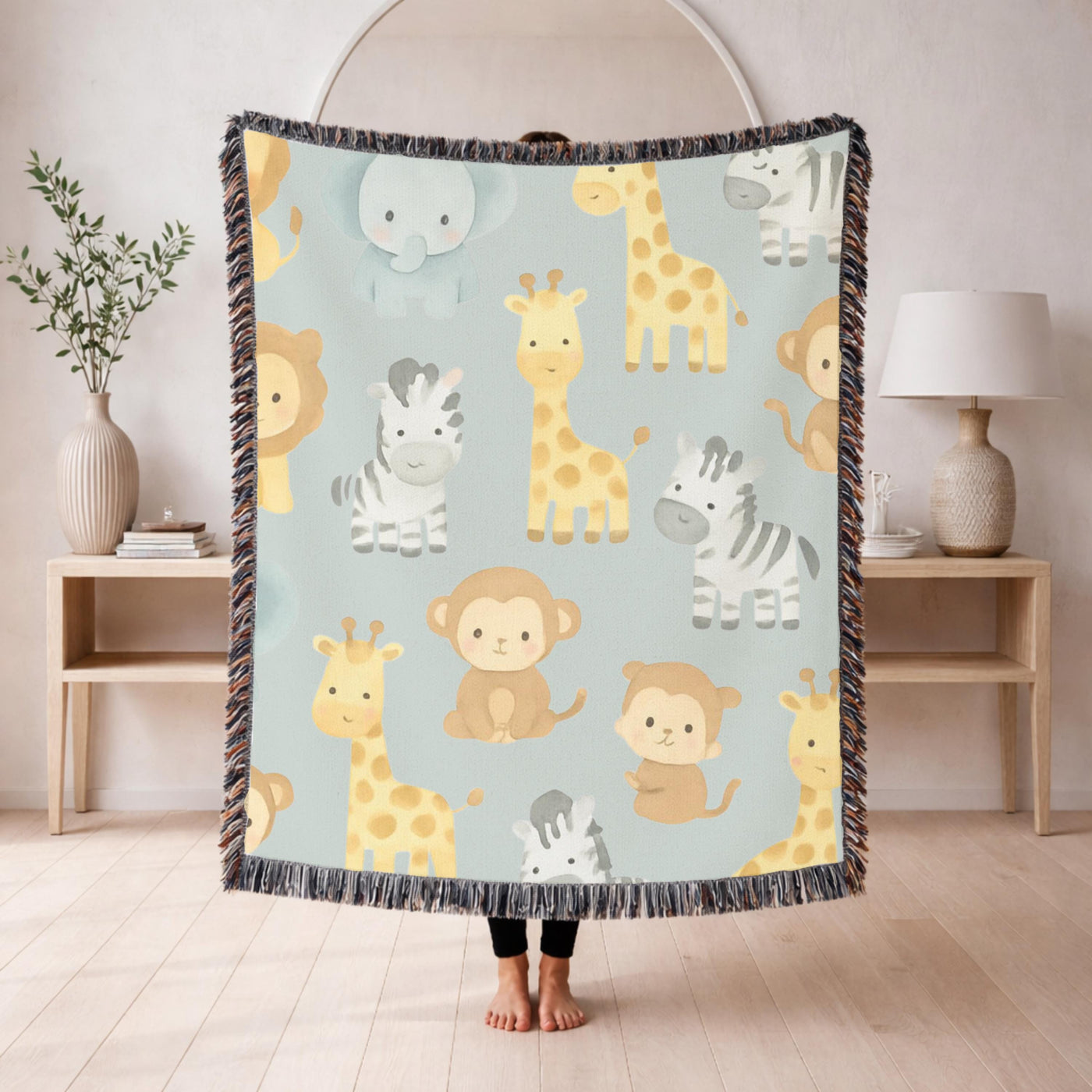 Fringed woven blanket in pale sage, printed with watercolour safari animals, giraffes, zebras, an elephant, a lion and monkeys, spaced evenly between small green leaves and sprigs.