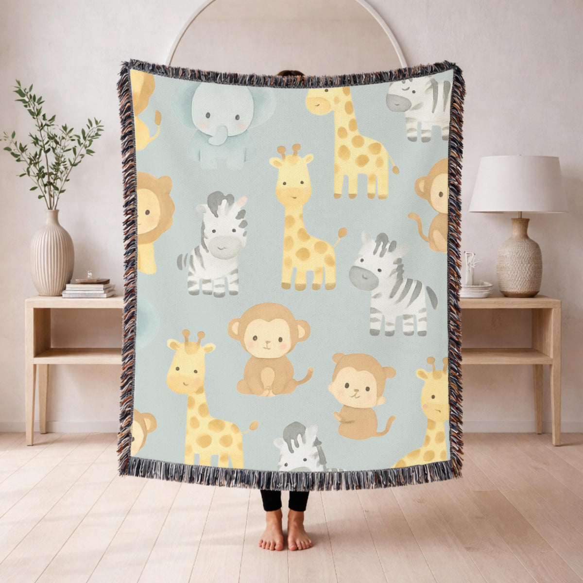 Fringed woven blanket in pale sage, printed with watercolour safari animals, giraffes, zebras, an elephant, a lion and monkeys, spaced evenly between small green leaves and sprigs.