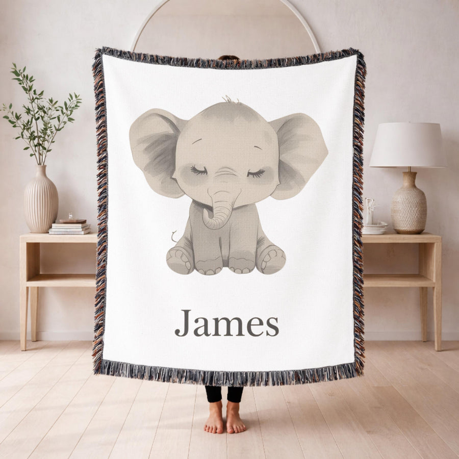 White fringed woven baby blanket draped over a wooden crib, printed with a large grey baby elephant sitting with its eyes closed and long lashes, and the name James in black serif below.