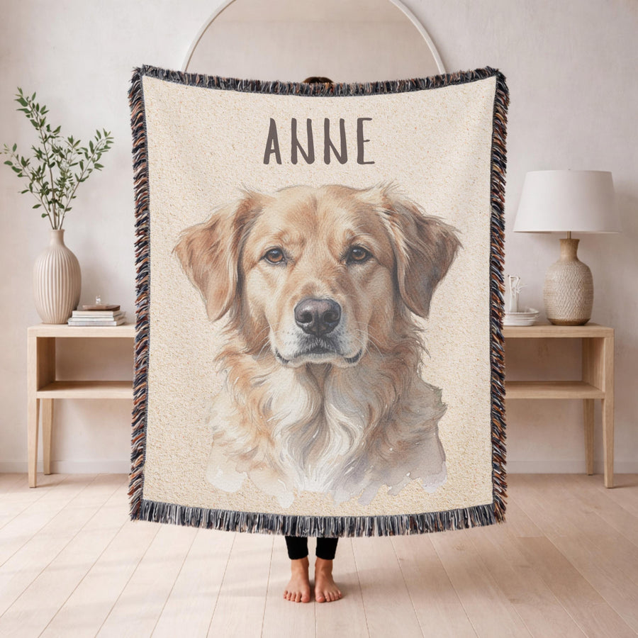 Cream fringed woven blanket held up in a living room, printed with a detailed watercolour portrait of a golden retriever, with the name ANNE in grey capital letters above it.