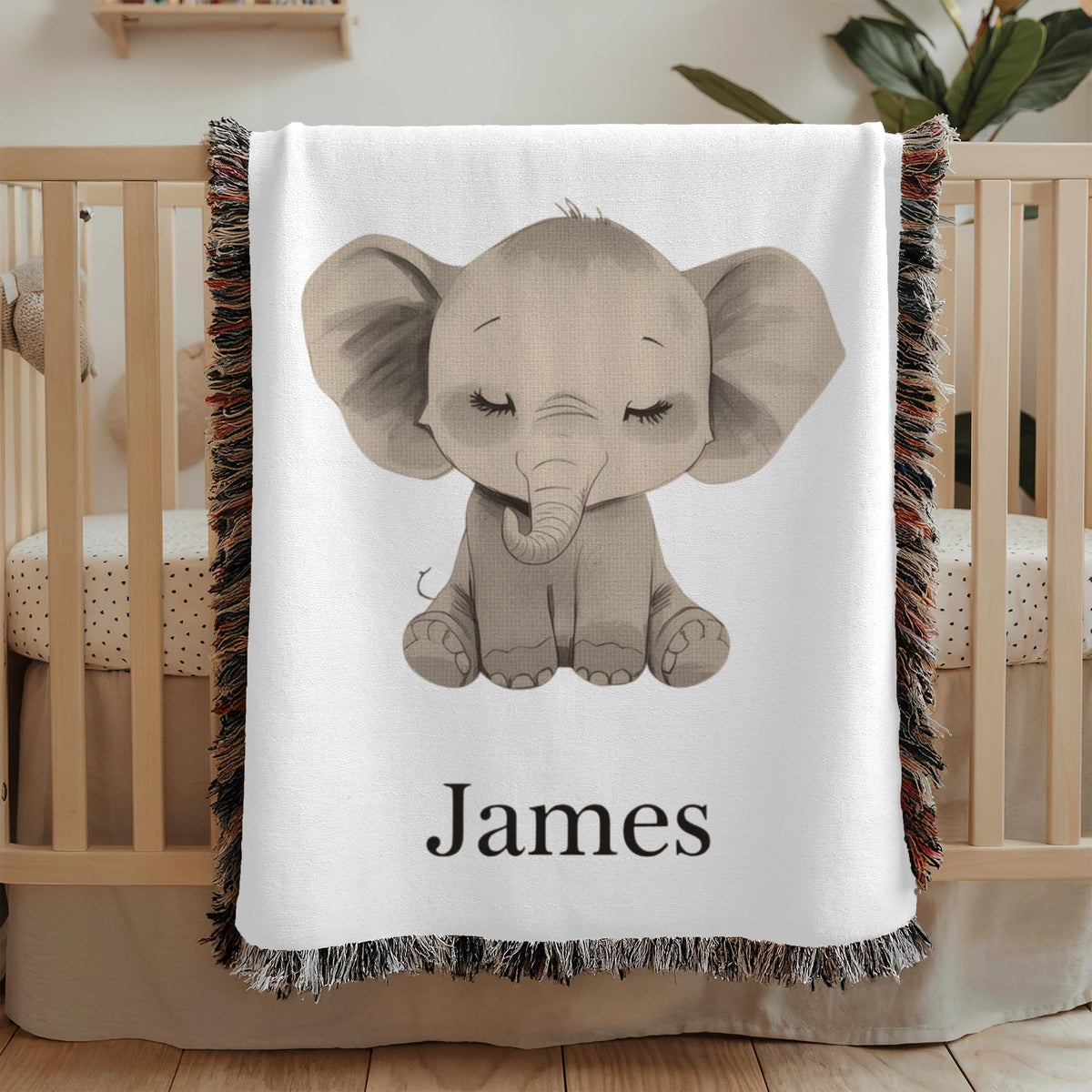White fringed woven baby blanket draped over a wooden crib, printed with a large grey baby elephant sitting with its eyes closed and long lashes, and the name James in black serif below.