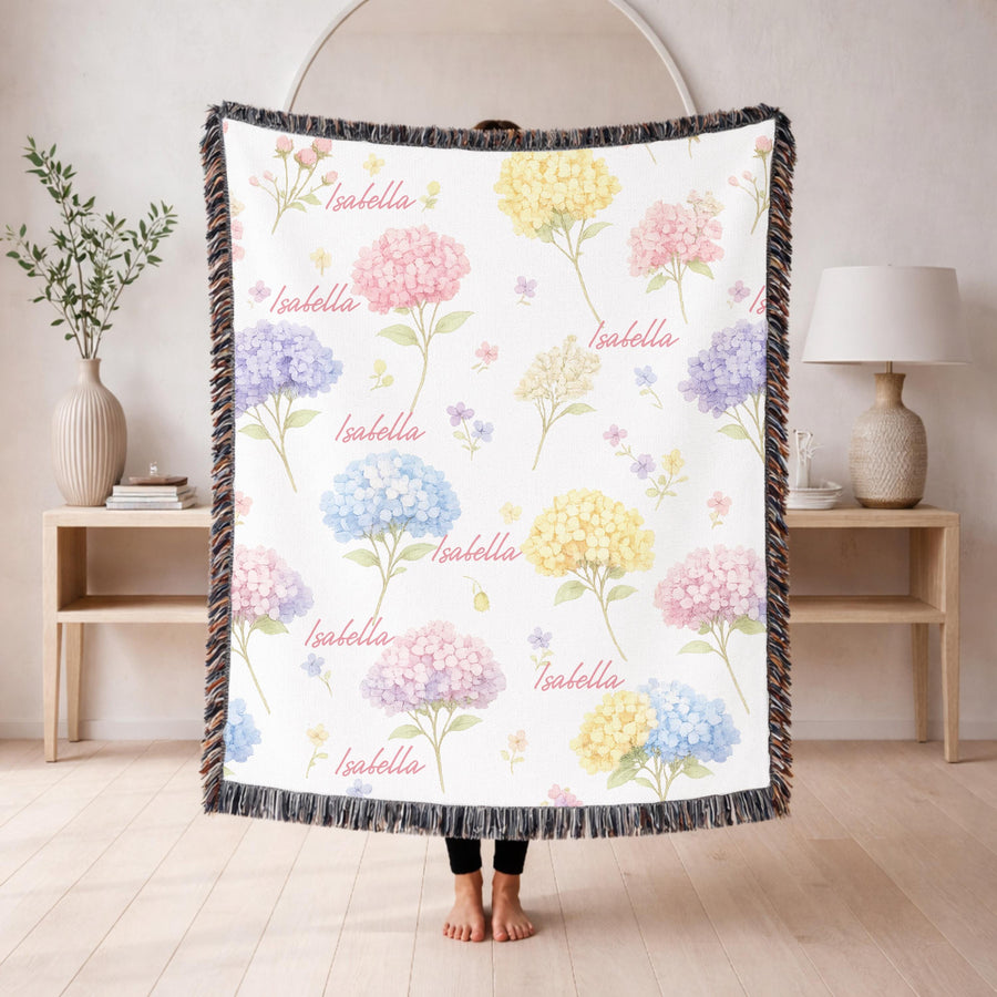 White fringed woven blanket over a wooden crib, scattered with pink, blue, yellow and purple watercolour hydrangeas, with the name Isabella repeated across it in pink script.