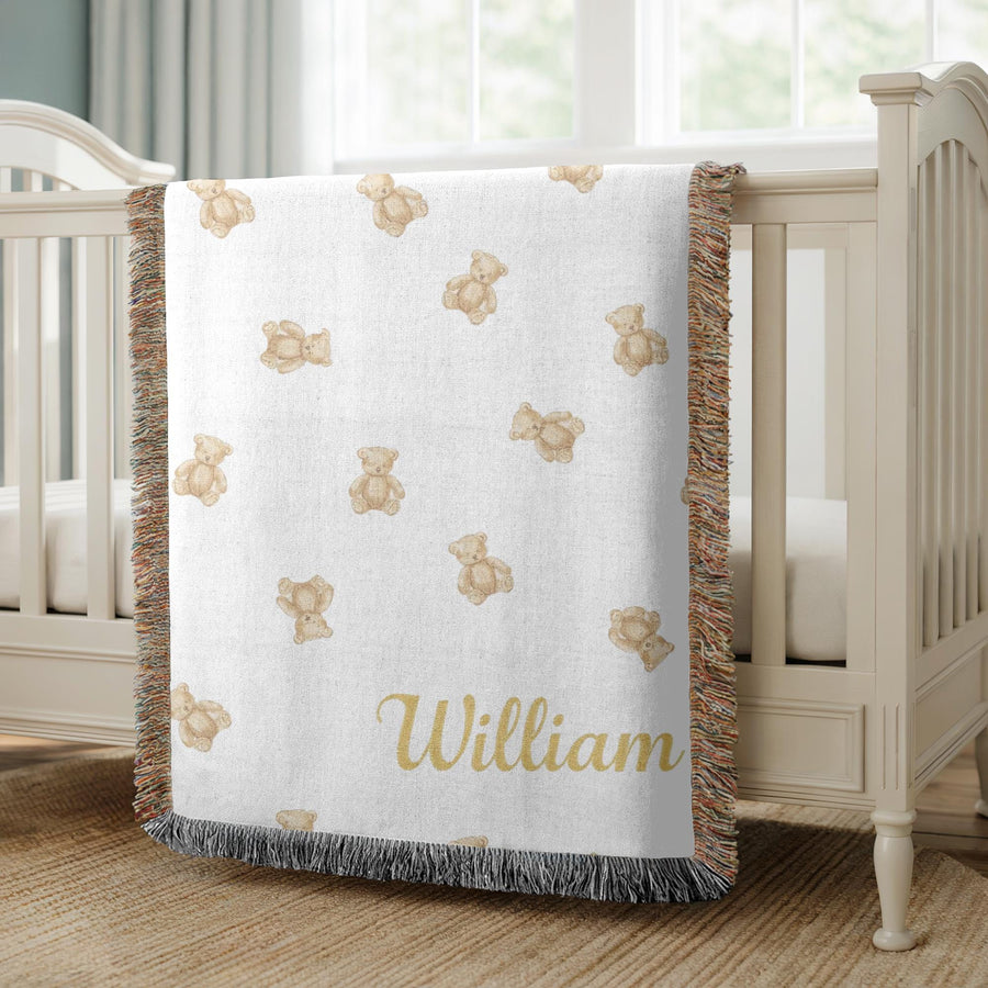 Cream fringed woven baby blanket over a nursery crib, scattered with small tan watercolour teddy bears and finished with the name William in gold script across the lower right corner.